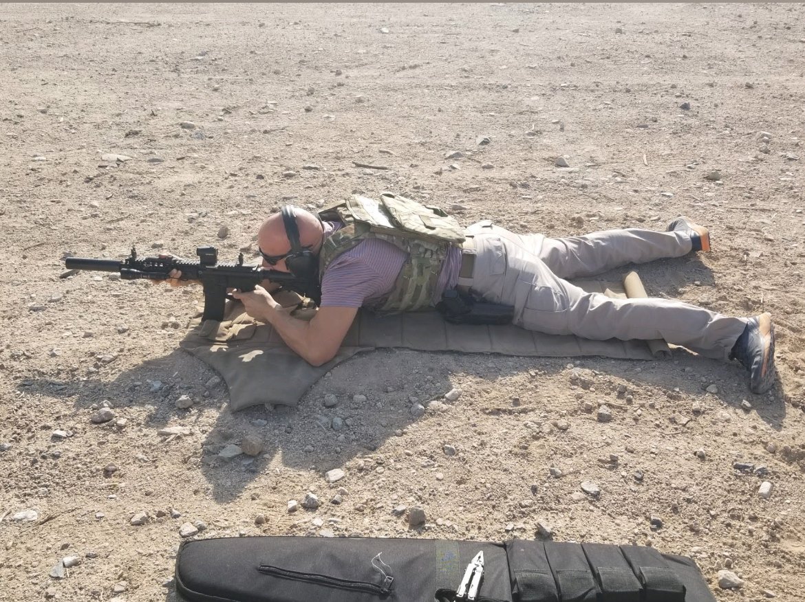 A man is lying prone on the ground aiming a rifle, wearing camouflage tactical gear, sunglasses, and ear protection in a desert environment.